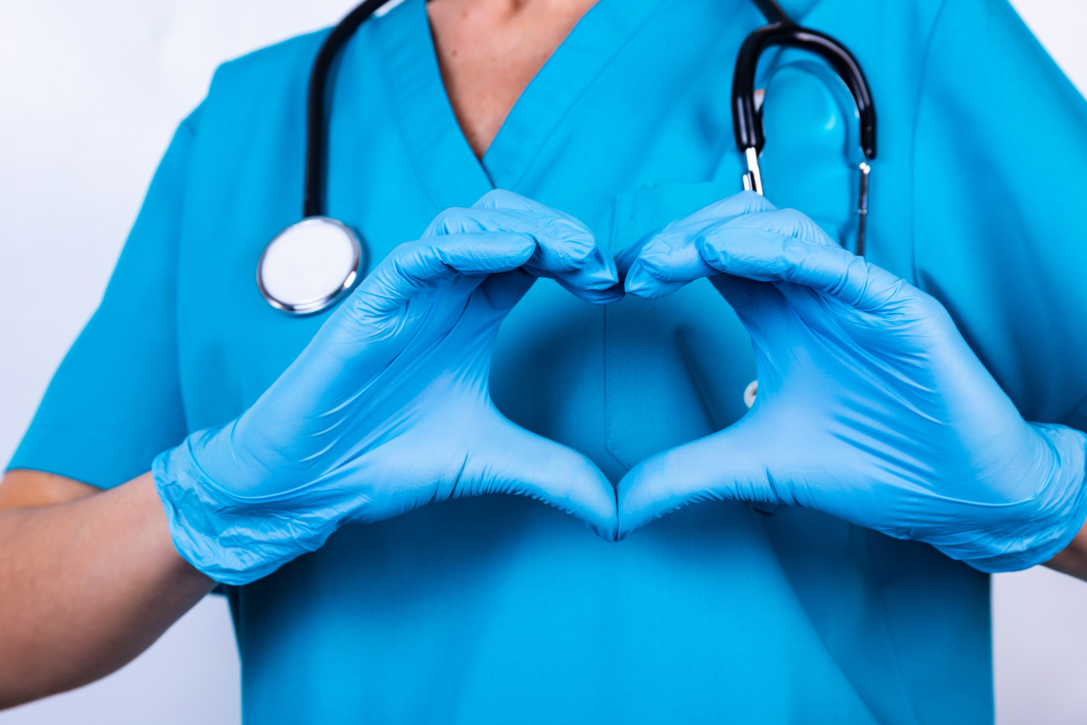 A doctor wearing hospital scrubs and a stethoscope forms a heart shape with their gloved hands over their heart.