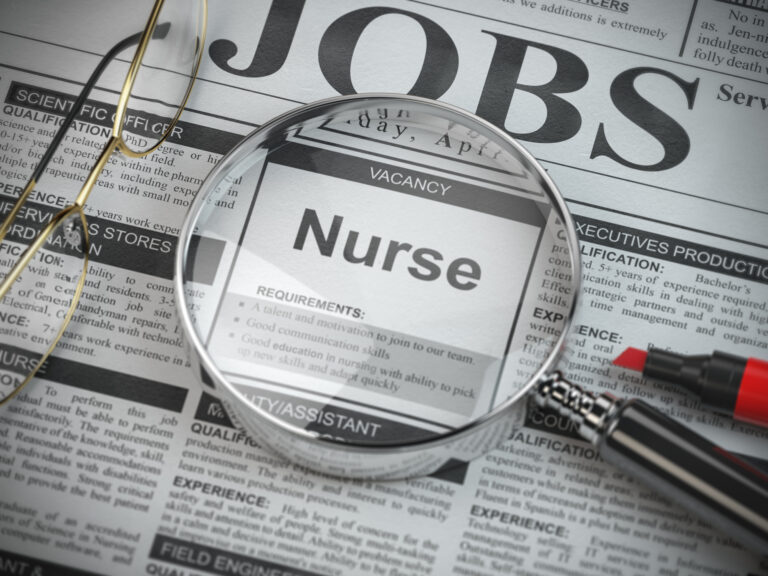 A magnifying glass lays on a newspaper's jobs page, focusing on the word "Nurse."
