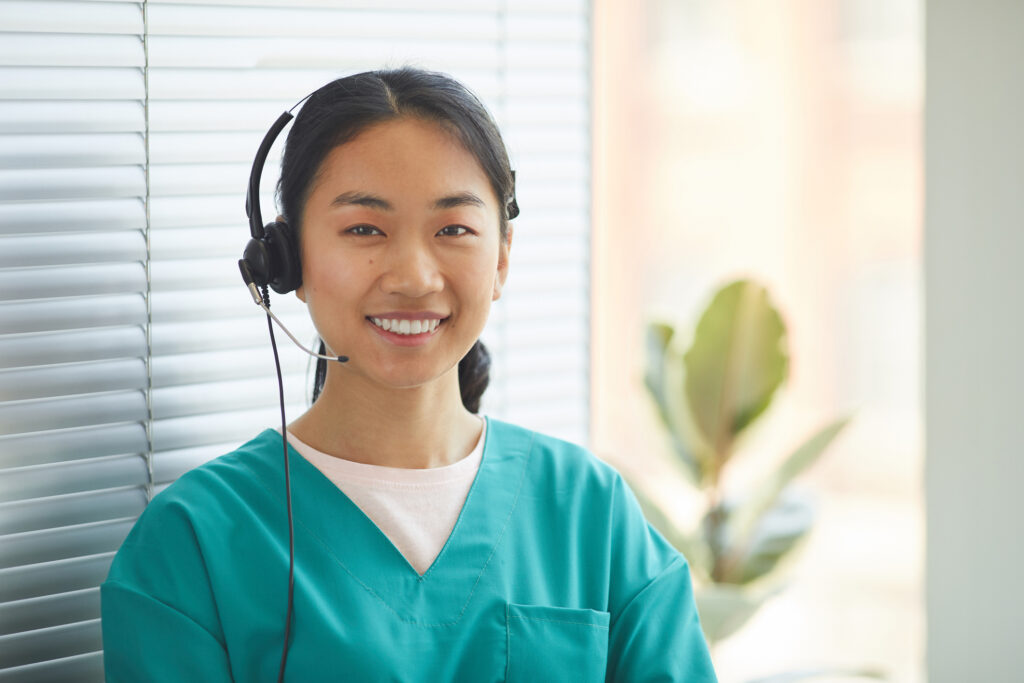 A registered nurse with a headset is standing and smiling in her office while performing telephone nurse triage.