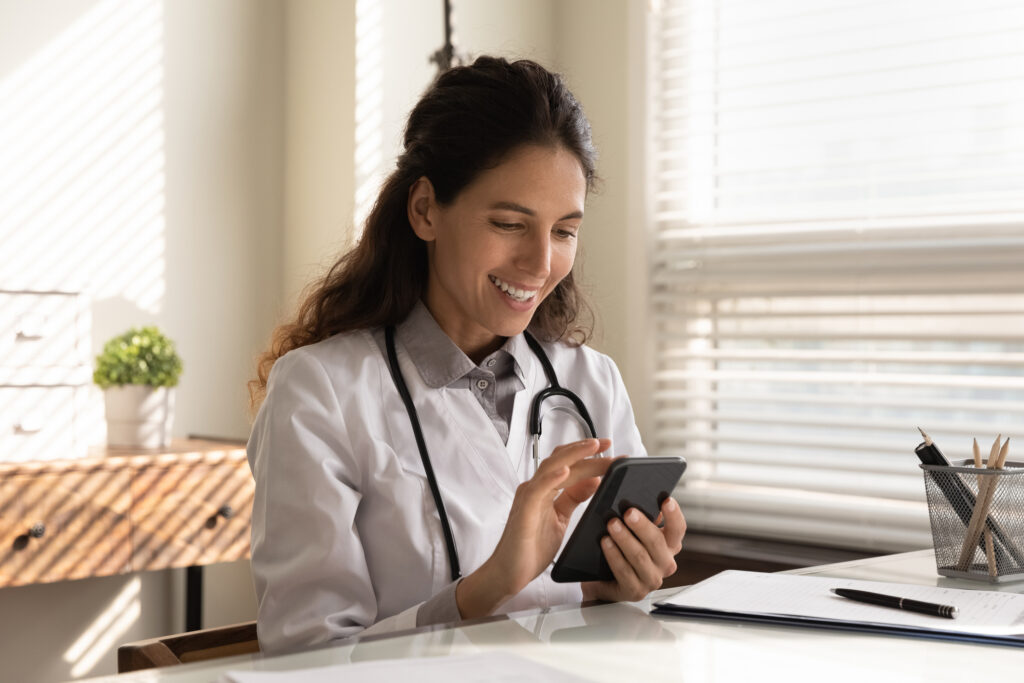 TriageLogic Secure Texting Young female doctor wearing a white lab coat and stethoscope sits at her office desk and smiles while checking her smartphone.