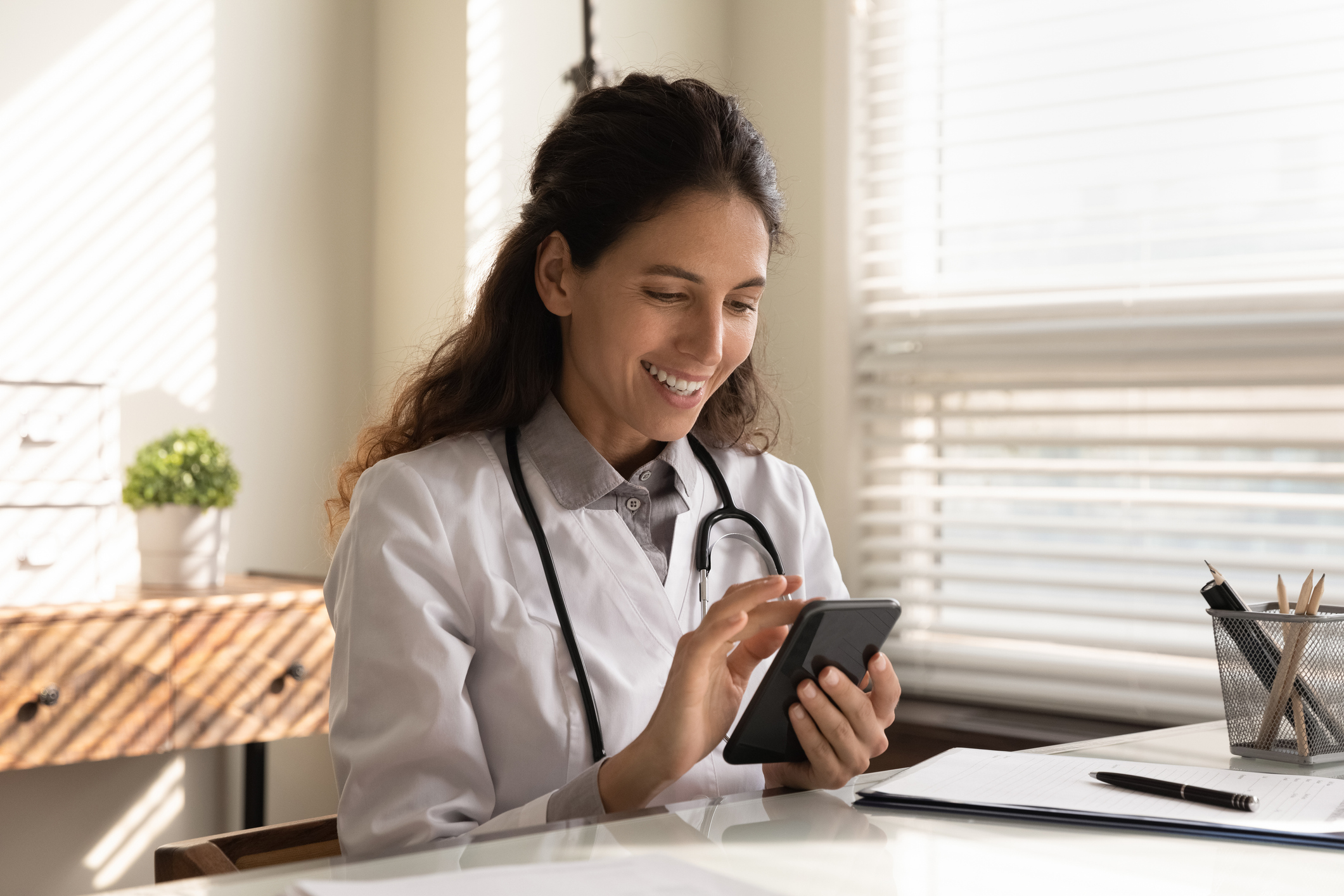 Young female doctor wearing a white lab coat and stethoscope sits at her office desk and smiles while checking her smartphone.