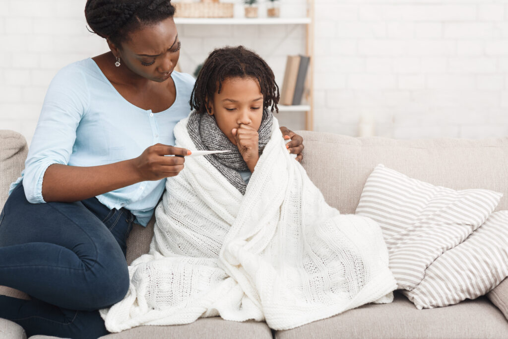 A mother comforts her child while sitting together on a couch. The mother checks a thermometer while the child is wrapped in a blanket and holds their hand to their mouth as they cough.