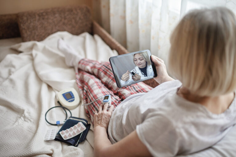 An older female patient lies in bed and uses a remote patient monitoring (RPM) device to record her vitals and share them with her provider during a telehealth call on her tablet.