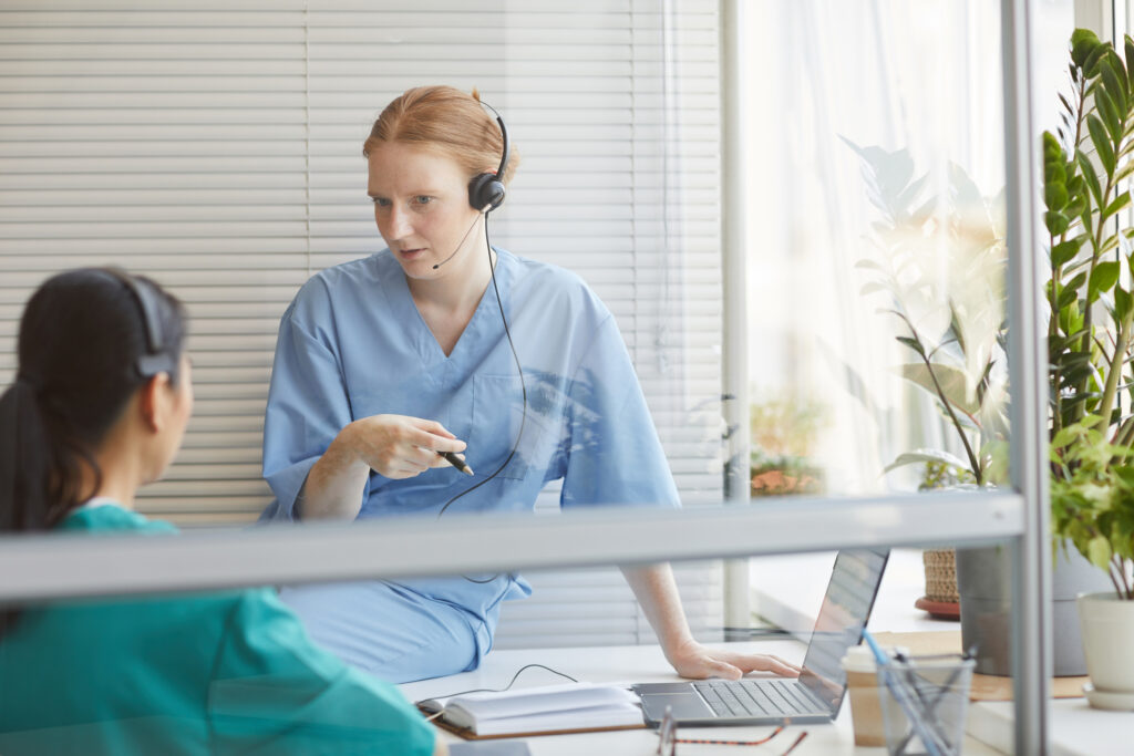 Two triage nurses wearing headsets discuss training beside an office window in a call center.