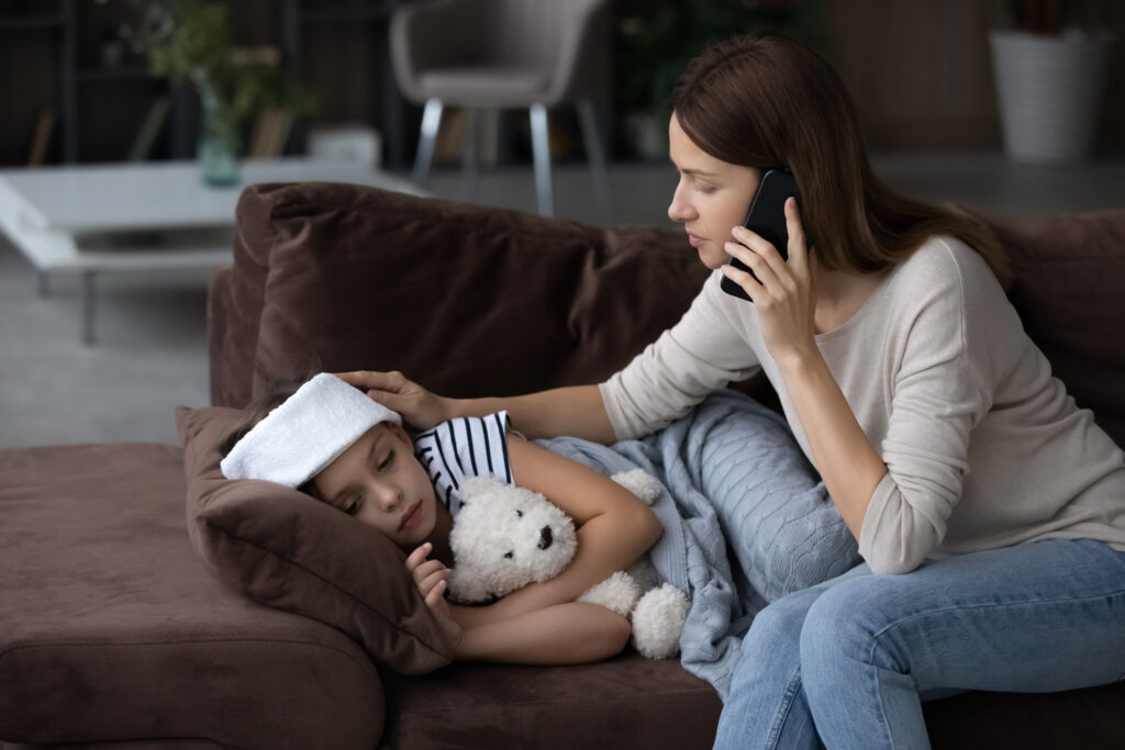 A mother comforts her daughter who lies on a couch with her teddy bear. The mother is on the phone with her pediatrician regarding the child's fever.