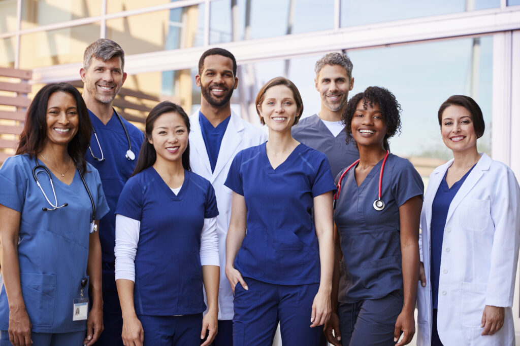 A diverse team of smiling nurses and physicians stands outside a hospital.