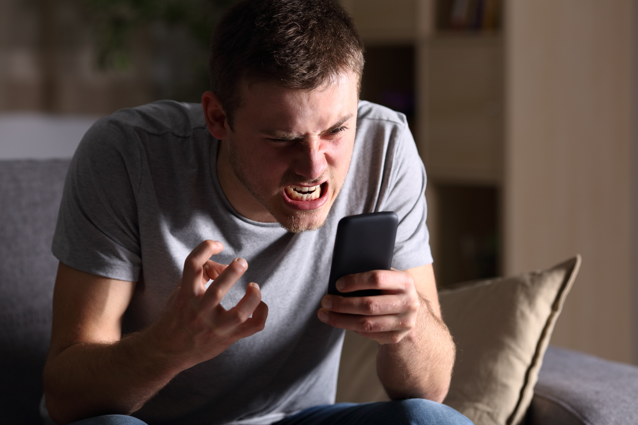 An angry patient caller sits on his couch at home and grimaces while yelling into a mobile phone in his hand.