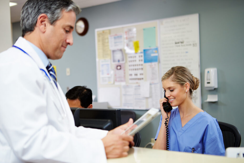 A nurse at a nurse's station in a hospital calls a patient back from a note they received from medical answering services, while a doctor reviews additional information beside her.