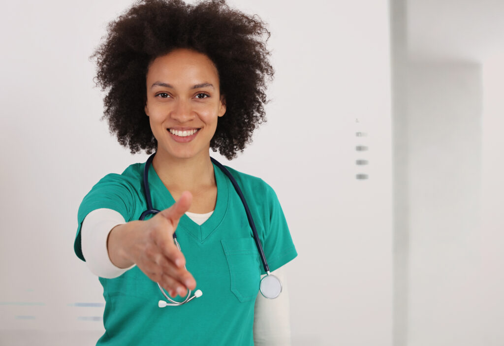A nurse in scrubs wearing a stethoscope holds out her hand to shake and welcome the viewer.