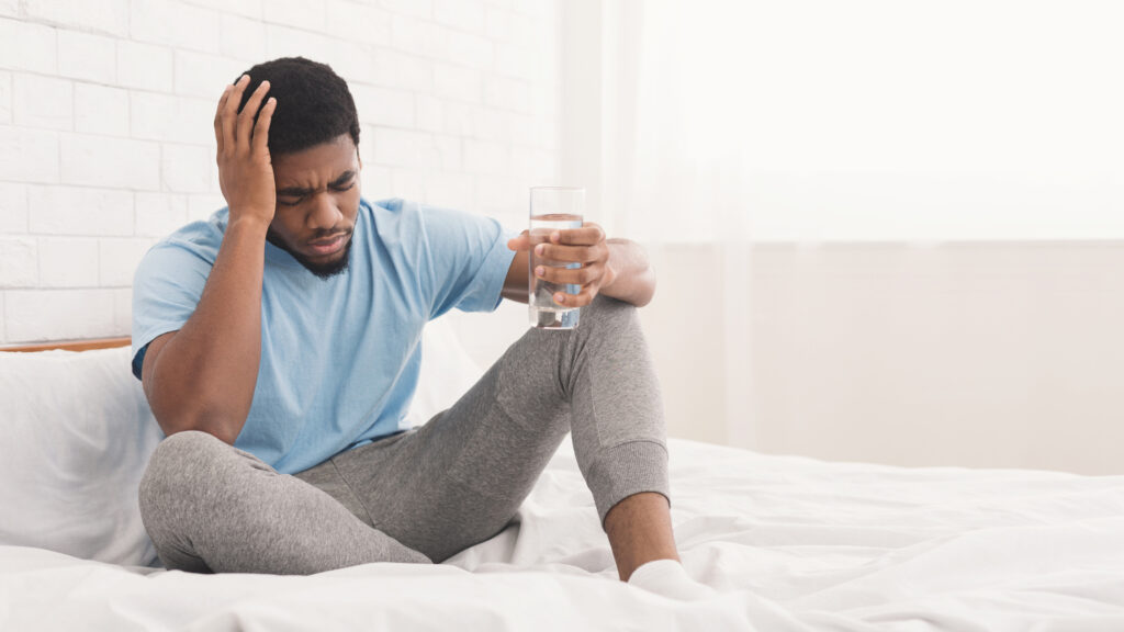 Dehydrated patients concept: a man sits upright in bed, holds a glass of water, and rests his head against his other hand.