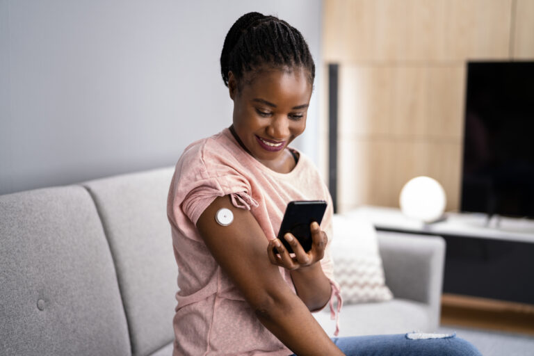 A patient uses a smartphone to check her glucose levels measured by an RPM device attached to her shoulder as part of type 1 diabetes management.