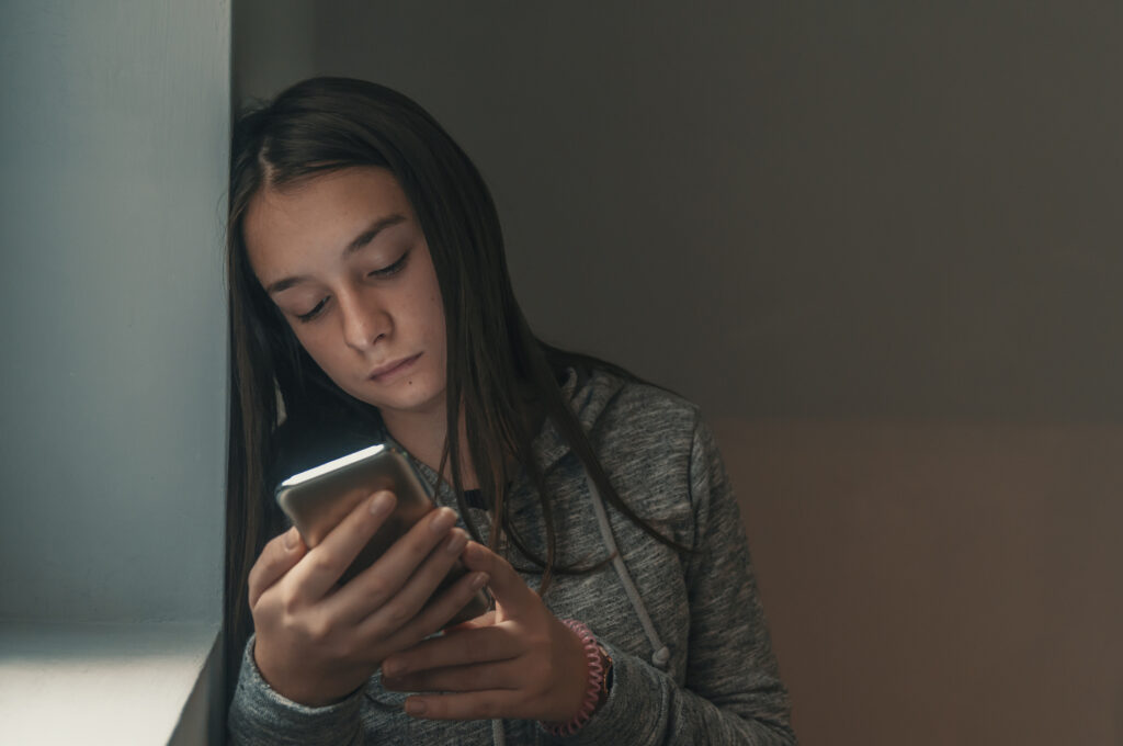 Student behavioral health outcomes concept: a sad student leaning against a window uses her smartphone to call her college's nurse triage line.