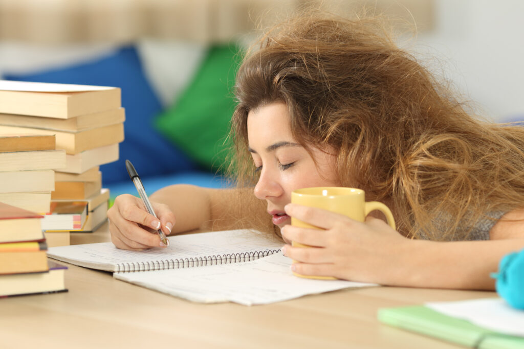 A sick student leans their chin against their notebook while writing and cradling a mug.