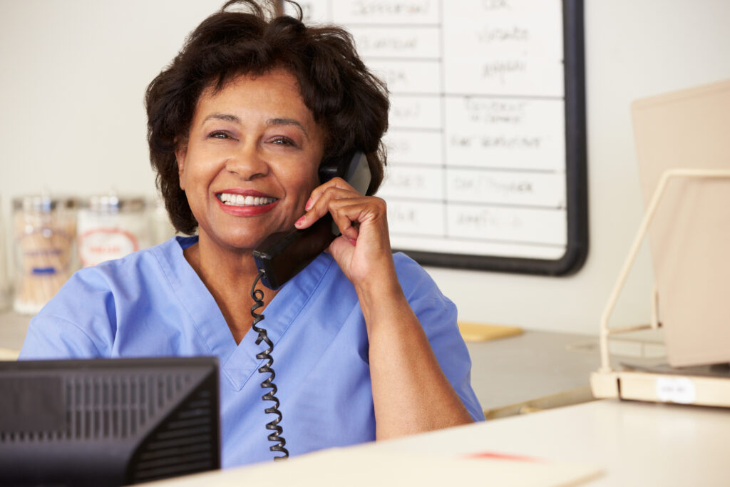 A triage nurse smiles while at her workstation giving care advice for patients over the phone.