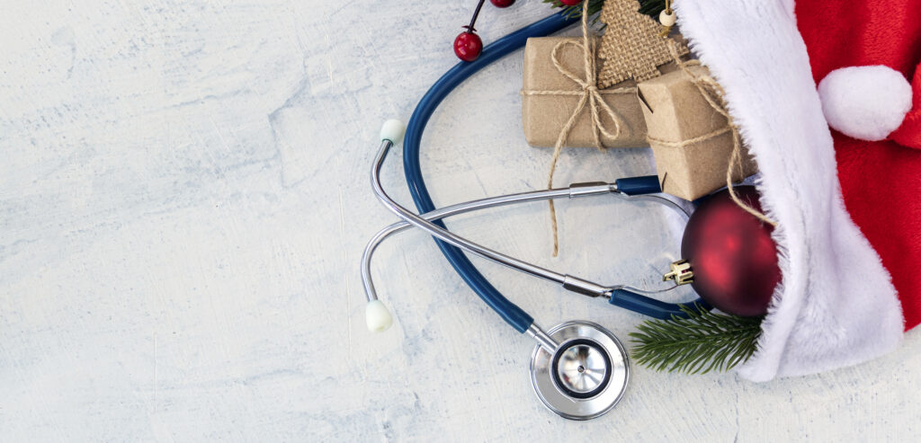 Concept for healthy triage nurses during the holidays: a stethoscope is packed into a Santa hat already filled with presents, an ornament, a red berry, and a pine tree branch.