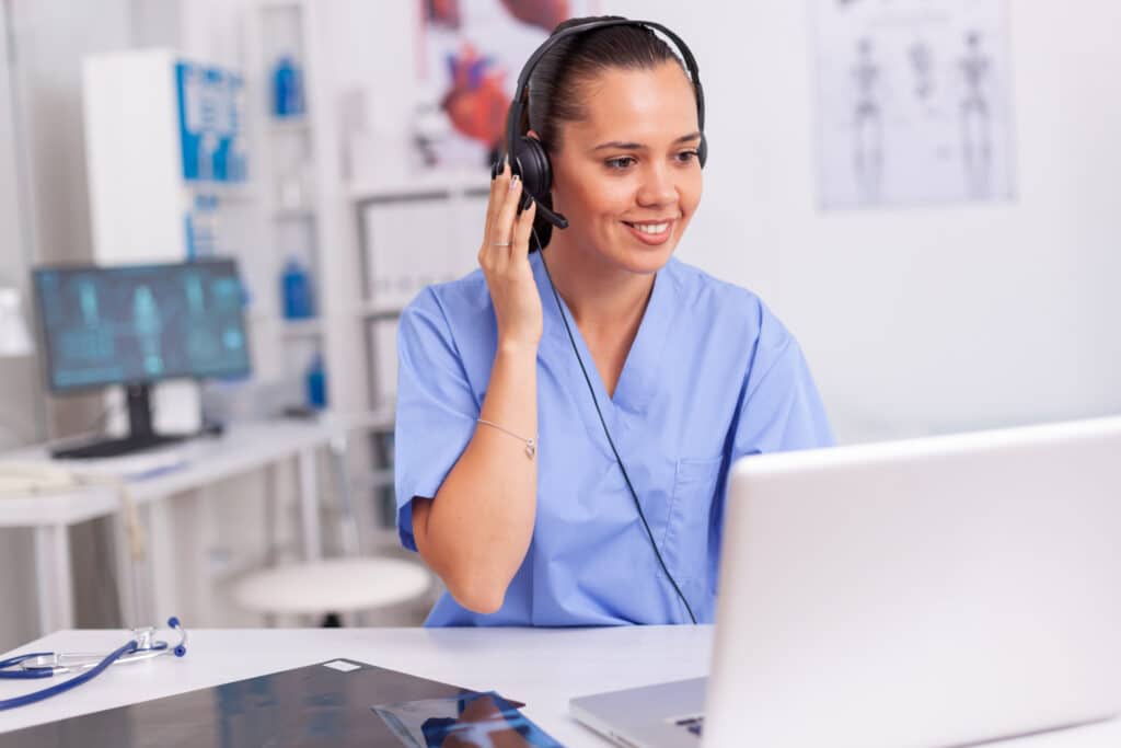 A triage nurse sitting in front of a laptop in a medical office uses a headset to talk with a patient about their symptoms.