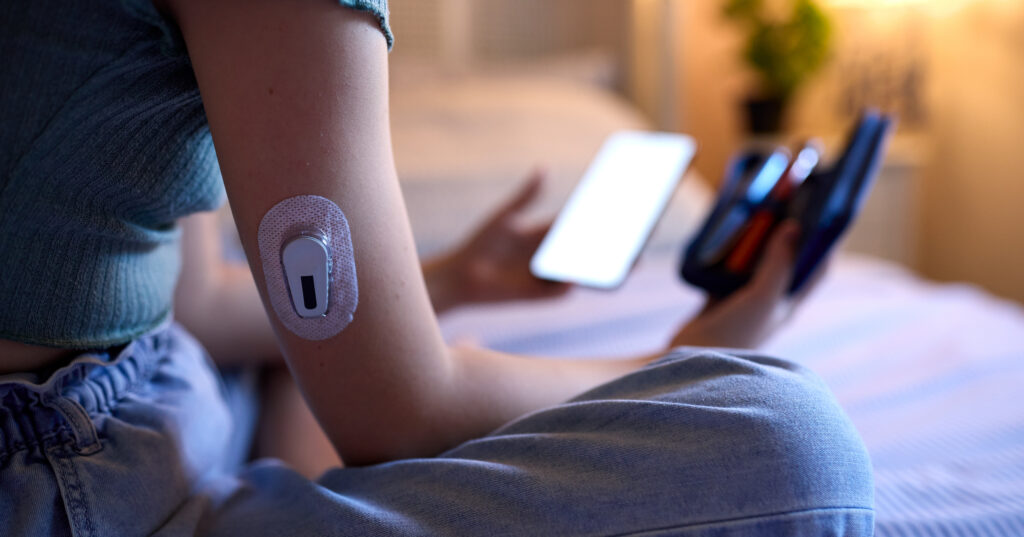 A diabetic patient sits on a bed checking their blood glucose level using a wearable sensor as part of a remote patient monitoring program.