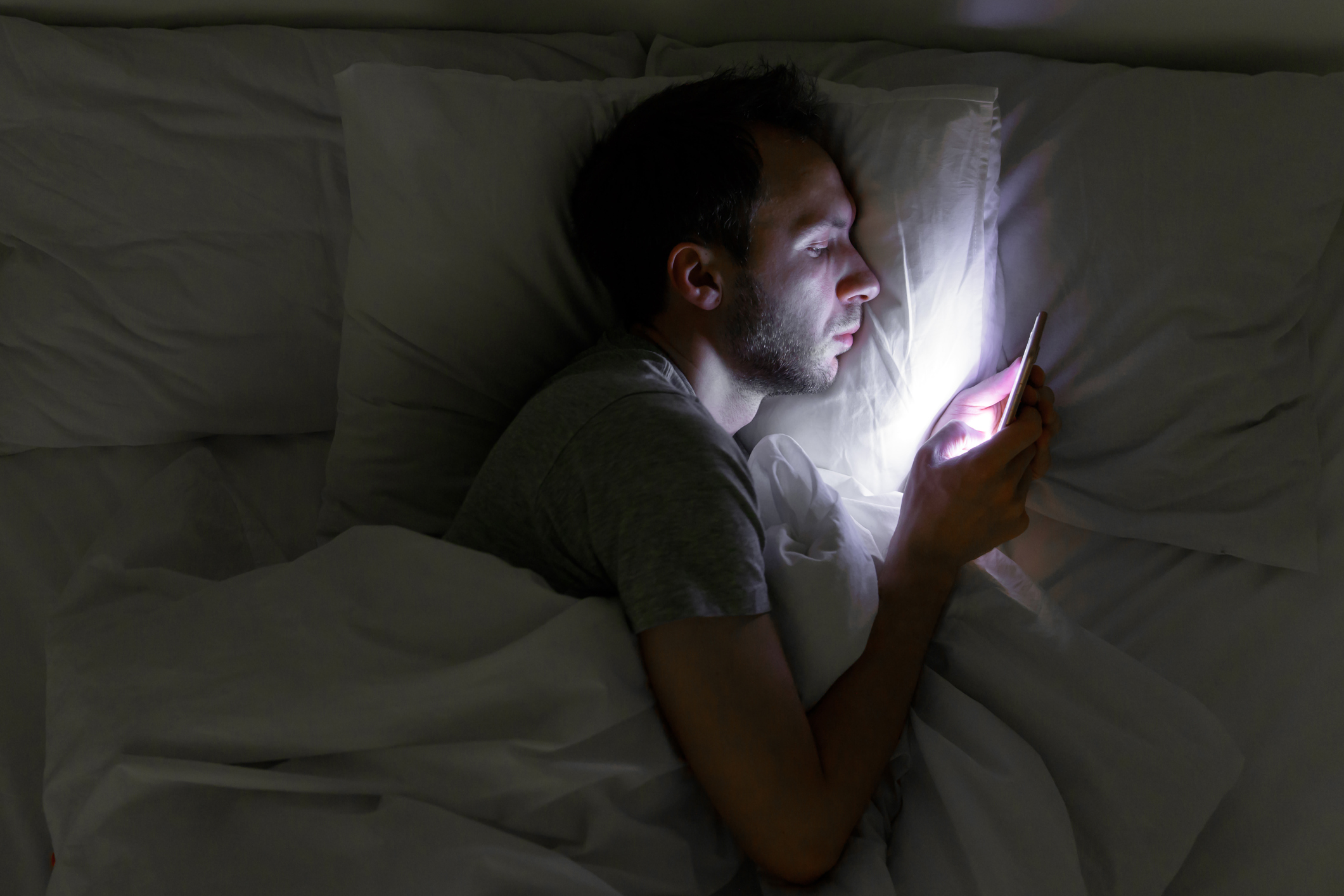 A patient lying in bed in the dark at home uses their smartphone to call after-hours nurse triage services.