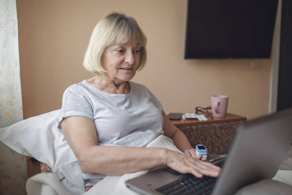 An older patient rests upright in bed while talking with her caregiver through her laptop. The RPM device she wears to monitor her vitals can not only improve her care, but also boost home healthcare revenue.