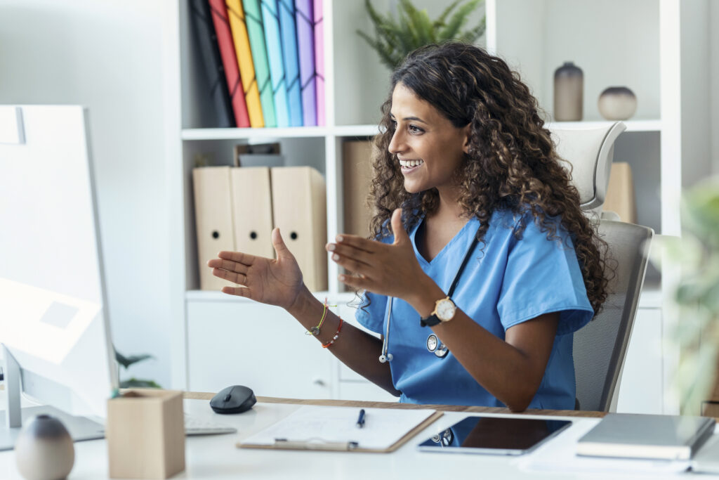 Telehealth nursing concept: a nurse smiles and gestures with her hands while talking with a patient through video conferencing on her computer.