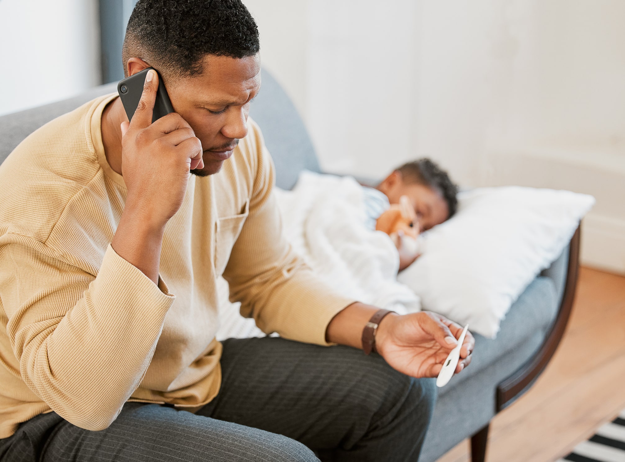 A parent sitting on a couch with their sick child at home calls pediatric telephone triage while checking their child's temperature.