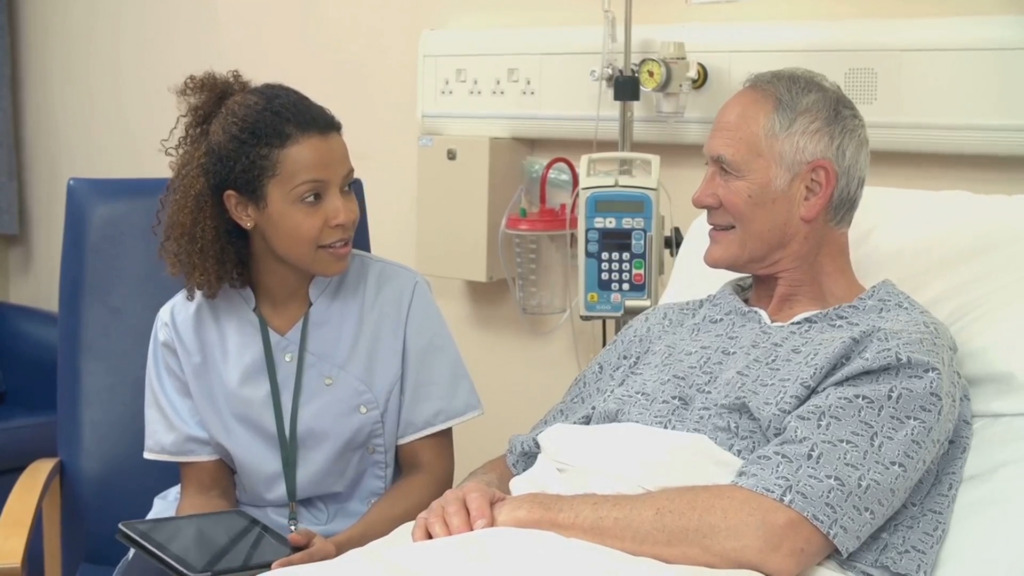 Nurse triage software benefits concept: a nurse sits at a patient's bedside explaining their health information and recording it on a tablet.