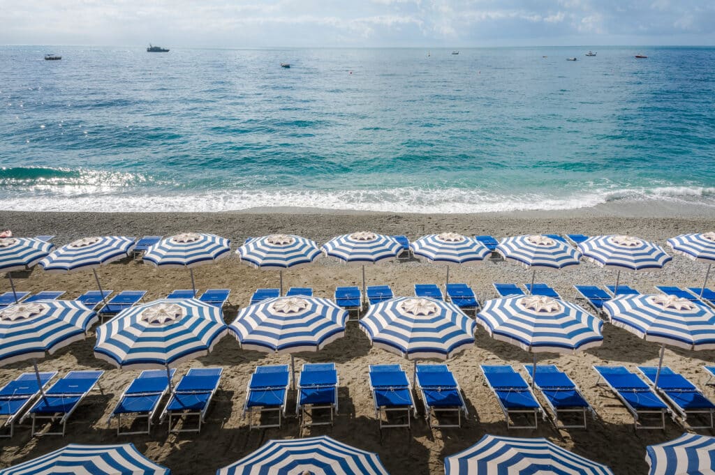 Several rows of beach chairs and umbrellas are arranged along the shoreline at midday and high tide.