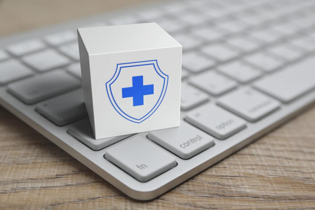 A block with a blue medical cross and shield is set on a keyboard, representing data security in remote patient monitoring.