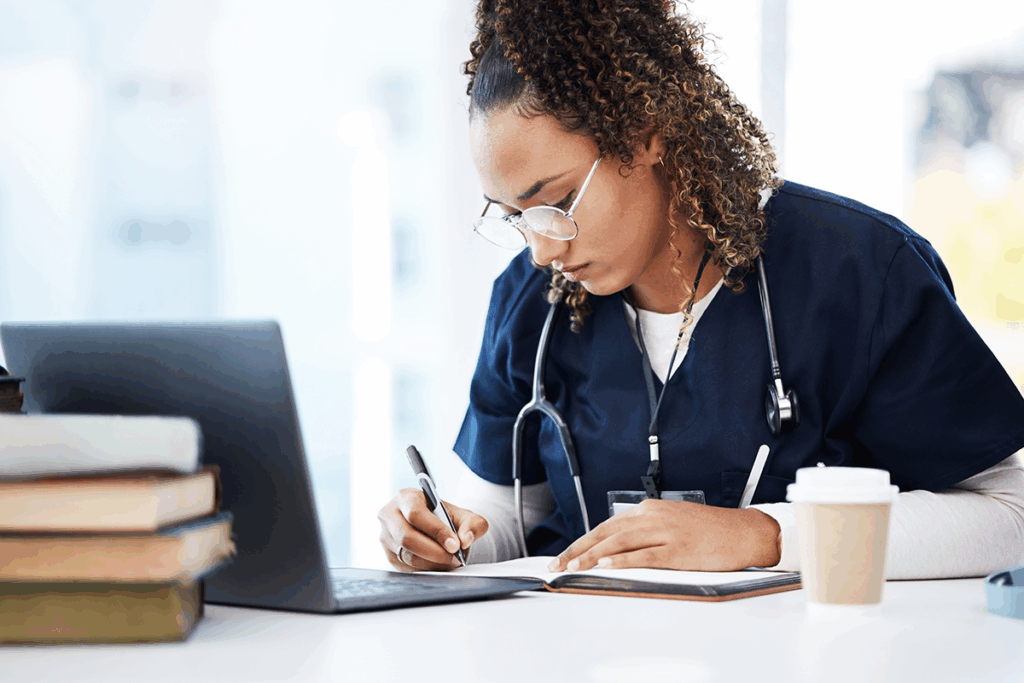 A triage nurse engages in continued education by writing information in her notebook and reviewing policies on a laptop.