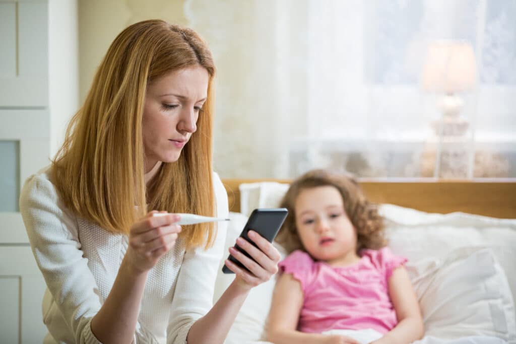 A parent uses her smartphone to access automated medical answering services for her sick daughter, who rests in bed.