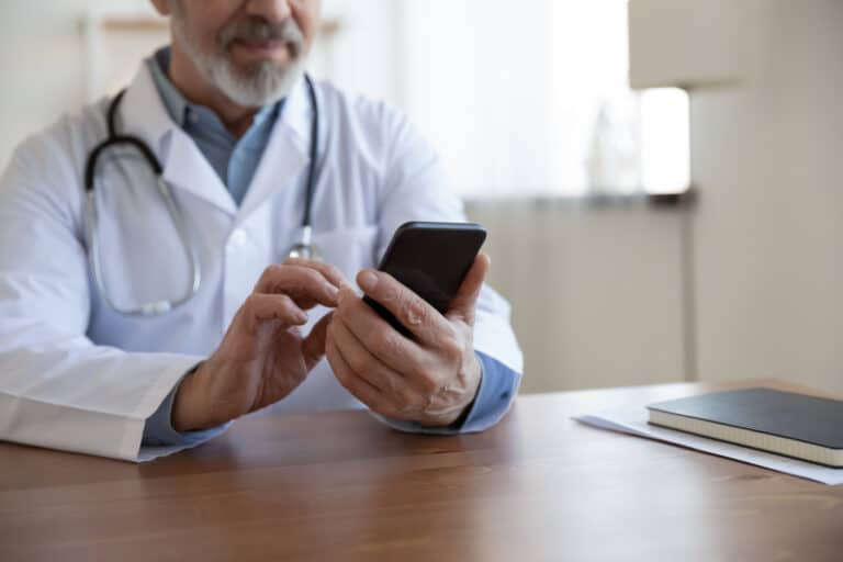 A physician sitting at a desk uses a secure messaging system on his smartphone to communicate with a triage nurse.