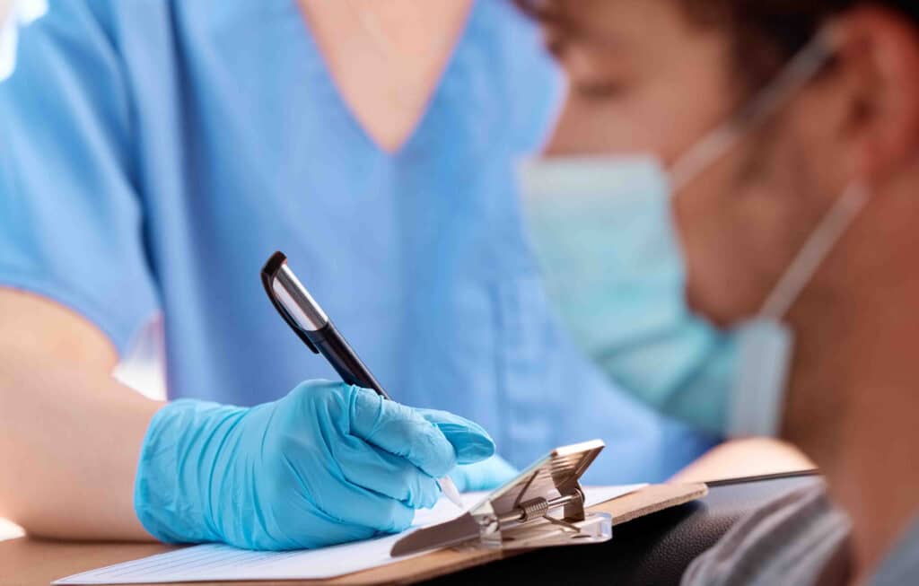 A nurse fills out a form on a clipboard while standing next to a doctor wearing a procedural mask.