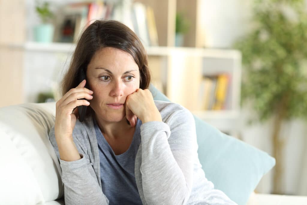 A female patient with a pensive expression sits on her couch at home, waiting on hold over the phone with hospital medical intake.