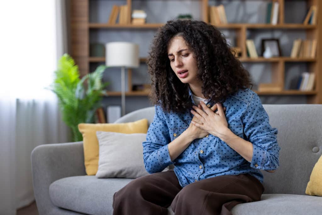 Panic attack vs heart problem symptoms represented by a woman holding her hands to her chest and experiencing heavy discomfort while sitting on a couch at home.