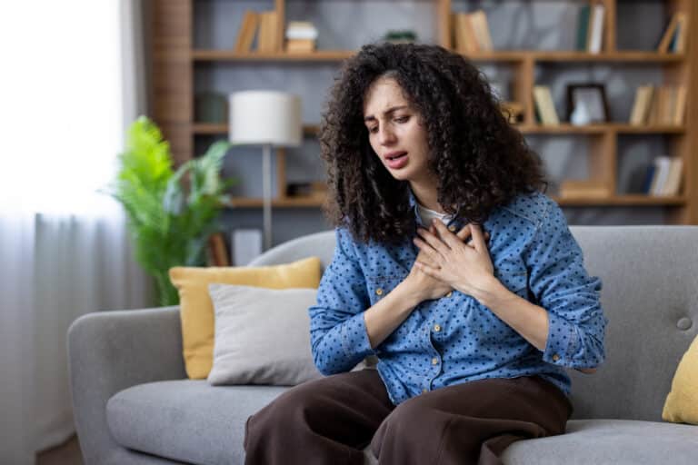 Panic attack vs heart problem symptoms represented by a woman holding her hands to her chest and experiencing heavy discomfort while sitting on a couch at home.