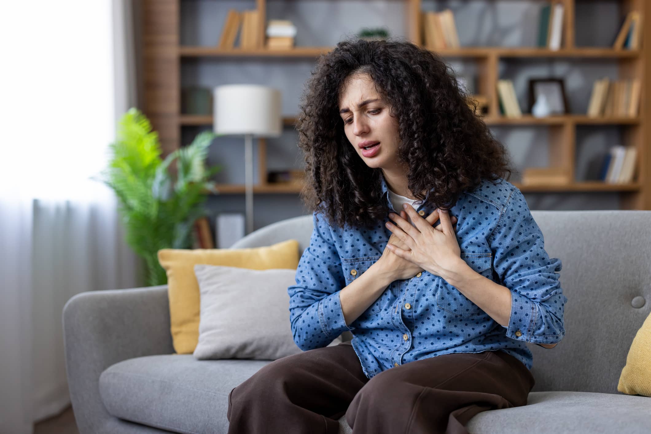 Panic attack vs heart problem symptoms represented by a woman holding her hands to her chest and experiencing heavy discomfort while sitting on a couch at home.