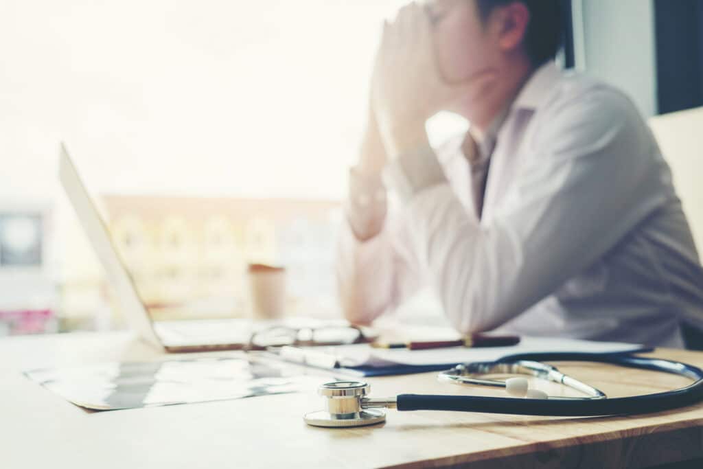 A tired physician sits at their desk and rests their head in their hands, illustrating challenges in medical call center workflow management.