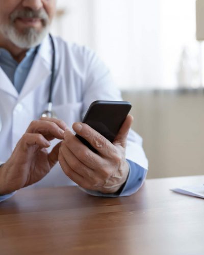 A physician sitting at a desk uses a secure messaging system on his smartphone to communicate with a triage nurse.
