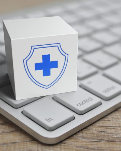A block with a blue medical cross and shield is set on a keyboard, representing data security in remote patient monitoring.