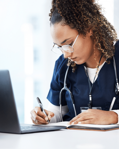 A triage nurse engages in continued education by writing information in her notebook and reviewing policies on a laptop.