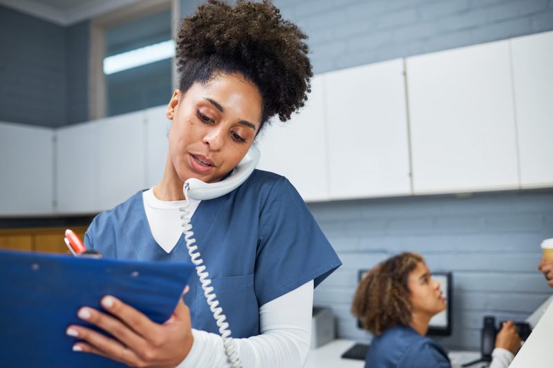 A triage nurse fills out information on a clipboard while talking with a patient over the phone.