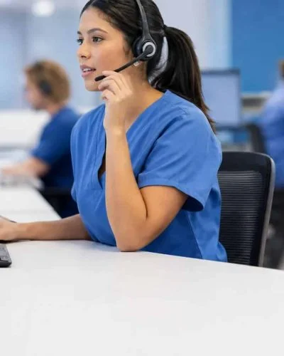 Registered nurse at a triage call center workstation reviewing patient information on dual monitors while speaking via headset.